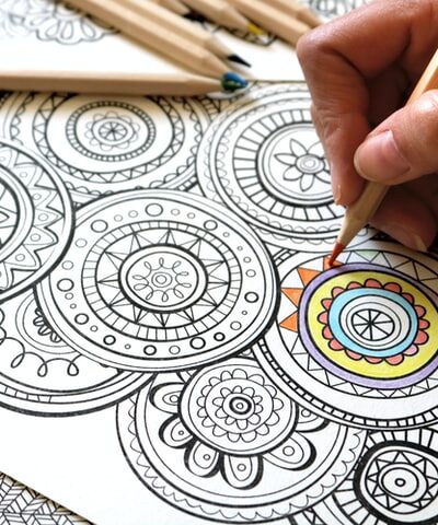 a womans hand holds a colored pencil while coloring a mandala coloring page