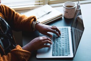 womans hands typing on a laptop at a desk with coffee and a notebook