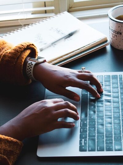 womans hands typing on a laptop at a desk with coffee and a notebook