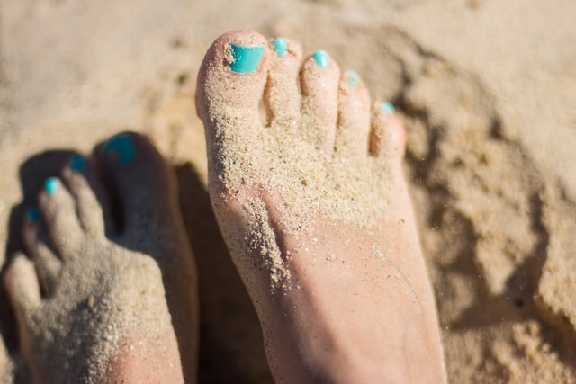 a womans feet on a beach with toes covered in sand wearing teal nail polish