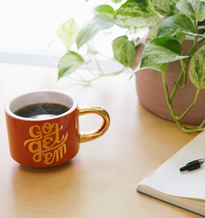 orange coffee cup that says go get em next to a notebook, pen, and plant on a table