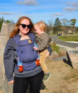 woman holding a toddler and wearing a baby in a carrier outdoors